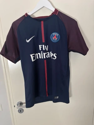 PSG matchtröja Nike blå vinröd - Snygg PSG matchtröja från Nike i mörkblått med vinröda ärmar och detaljer. Tröjan har klubbmärke och Fly Emirates-tryck på bröstet samt vit Nike-logga. Tillverkad i lätt och ventilerande material, perfekt för fotboll eller träning.