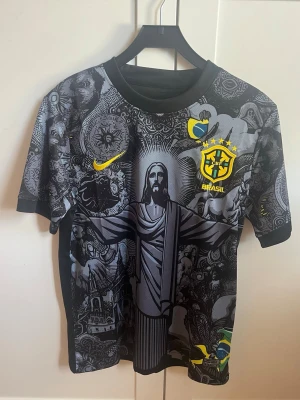 Nike Brasil svart fotbollströja - Unik svart fotbollströja från Nike med Brasil-logga och gult tryck. Tröjan har ett detaljerat grafiskt mönster med Kristusstatyn och brasilianska symboler i gråskala. Rund halsringning och korta ärmar. Perfekt för dig som vill sticka ut på planen.