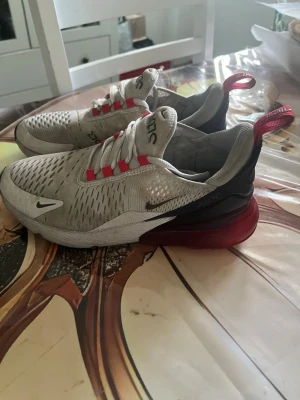 Nike 270 vit röda  - Nike Air Max 270 sneakers i vitt meshmaterial med svarta och röda detaljer. Skorna har en stor synlig Air-enhet i hälen, röda öglor och svarta partier bak. Swoosh-logga på sidan och ventilerande ovandel. Perfekta för dig som gillar sportig och modern stil.