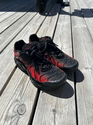 Nike Air Max Plus svart/röd - Säljer ett par Nike Air Max Plus sneakers i svart och rött med ikoniska vågmönster på ovandelen. Skorna har en chunky sula och meshmaterial för bra andningsförmåga. Klassisk Nike-logga på sidan och tunna svarta skosnören. Perfekta för dig som gillar streetwear.