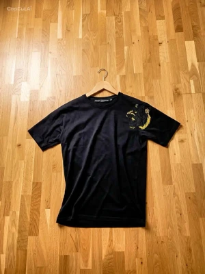 Svart Philipp Plein t-shirt med tiger tryck  - Svart premium tränings- och livsstilströja med exklusivt guldfärgat lejonmotiv som ger ett kraftfullt och stilrent intryck. Tröjan har en modern passform och är tillverkad i ett lätt och bekvämt material som passar lika bra till träning som till vardags. Den minimalistiska svarta designen kombinerad med den detaljerade guldloggan skapar en lyxig känsla och gör att tröjan sticker ut på ett elegant sätt.  Skick: Mycket fint skick utan synliga skador eller större slitage. Färg: Svart med gulddetalj