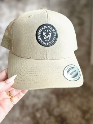 Beige keps Broken Society snapback HELT NY! - Snygg beige keps från Broken Society med svart och vitt runt märke framtill med döskalle och text. Klassisk snapback-modell med justerbar baksida och meshpaneler för extra ventilation. Perfekt för en avslappnad streetwear-look. - OANVÄND!