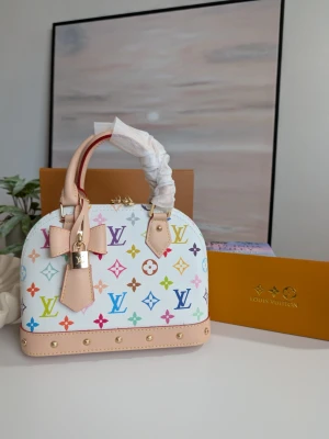 Iouis Vuitton BB Murakami Edition 🌸 - En dröm i färg och form Denna ljuvliga handväska utstrålar en tidlös charm med sin mjuka, kupolformade silhuett som påminner om klassisk elegans. Den står stadigt på en vacker bas, redo att bära dina mest värdefulla ägodelar genom livets alla små och stora ögonblick.Väskan omfamnas av två handtag i naturligt, ljusbeige läder som med tiden kommer att bära spår av dina resor och äventyr.