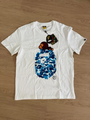 Vit BAPE t-shirt med blått camomönster - Säljer en vit t-shirt från BAPE med ikoniskt blått camomönster och stor apa-tryck på bröstet. Klassisk rund hals och korta ärmar. Märkets taggar sitter kvar och motivet är riktigt färgstarkt och unikt.