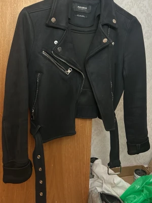 Svart jacka från Pull&Bear - Snygg svart jacka från Pull&Bear med klassisk bikerstil. Jackan har silvriga dragkedjor, axelklaffar och ett brett bälte i midjan. Kortare modell med detaljerade sömmar och tryckknappar på kragen. Perfekt för en edgy look.