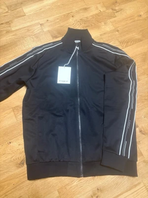 Svart ziptröja från Givenchy - Svart ziptröja från Givenchy med vita ränder längs ärmarna och diskret logga på ärmen. Tröjan har hög krage, muddar vid ärmslut och nederkant samt fickor framtill. Perfekt för en sportig och trendig look. Helt oanvänd , ny pris 13-14 tusen. 100% äkta köpt från Miinto.