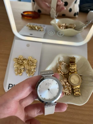 Vit klocka med stenar från ZONE - Stilren klocka från ZONE med vitt armband och silverfärgat ur. Urtavlan är enkel med små stenar som timmarkeringar och extra stenar på boetten. Armbandet är i syntetmaterial och har klassiskt spänne. Perfekt accessoar för dig som gillar en clean look.