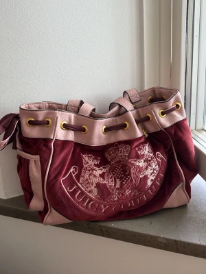Juicy Couture daydreamer - Juicy Couture daydreamer i vinrött och ljusrosa med stora guldfärgade öljetter och dragskostängning. Väskan har broderad logga framtill samt mönstrat innerfoder med innerficka i guld. Självklart äkta.