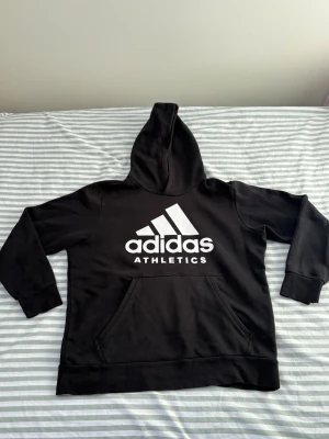 Svart Adidas-hoodie med dolt blixtlåsficka - Svart hoodie med vit Adidas-logotyp. Tröjan har en känguruficka framtill och en liten ficka med dragkedja inuti på höger sida. Huvan är enkel och tröjan har muddar i ärmslut och nederkant. Om du gillar den där baggy hoodien är den här perfekt för dig.