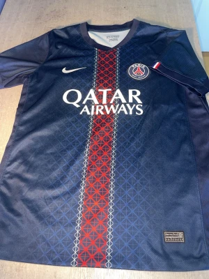 PSG matchtröja Nike blå/röd - Snygg PSG matchtröja från Nike i mörkblått med röd och vit rand i mitten. Tröjan har klubbmärke och Nike-logga på bröstet samt Qatar Airways som sponsortryck. Tillverkad i lätt och ventilerande material, perfekt för fotboll eller träning.