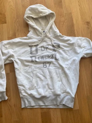 Vit hoodie med tryck och huva - Säljer en vit hoodie från Ralph lauren med stor magficka och tryck i grått framtill. Hoodien har dragsko i huvan och ribbade muddar vid ärmslut och nederkant. Perfekt för en avslappnad och chill stil.