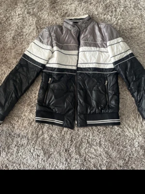 D&G bomberjacka i svart, vitt och grått - Snygg bomberjacka från Dolce & Gabbana i svart, vitt och grått med sportiga ränder. Jackan har dragkedja framtill, två fickor med dragkedja och ribbade muddar med ränder. Materialet är glansigt och quiltat för en modern look.