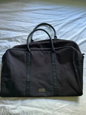 Svart weekendbag från Hugo Boss - Stilren svart weekendbag från Hugo Boss i tåligt tyg med detaljer och handtag i svart läderimitation. Väskan har en klassisk rektangulär form och metallbricka med logga framtill. Perfekt för resor när du vill ha både stil och funktion.