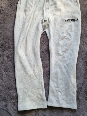 Grå Essentials mjukisbyxor - Snygga ljusgrå mjukisbyxor från Fear of God Essentials med tryckt logga på ena benet. Byxorna har dragsko i midjan och raka ben. Tillverkade i mjukt och bekvämt material, perfekta för chill dagar. Klassisk streetwear-look som funkar till det mesta.