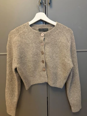 Beige cropped kofta från Arket - Mysig beige cropped kofta från Arket med rund halsringning och fyra träknappar framtill. Koftan är stickad i ett mjukt material och har långa ärmar samt ribbade muddar vid ärmslut och nederkant. Perfekt för lager på lager och enkel att styla.