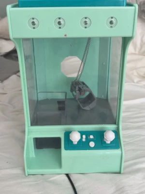 Mintgrön mini-claw machine - Säljer en mintgrön mini-claw machine i plast med två vita spakar och knappar på kontrollpanelen. Maskinen har genomskinliga sidor och en klo i metallkedja för att plocka upp småsaker. Perfekt för att ha hemma som rolig elektronikpryl.
