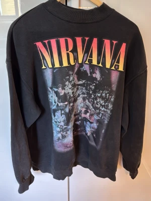 Nirvana Unplugged sweatshirt svart - Säljer en svart sweatshirt med stort Nirvana-tryck fram och bak. Framsidan har motiv från Unplugged-konserten och baksidan har låtlista från spelningen. Tröjan har rund hals, ribbade muddar och är gjord i mjukt bomullsmaterial. Perfekt för dig som älskar grunge och vill sticka ut.