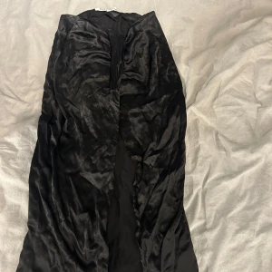 Svart långkjol från NA-KD - Säljer en svart långkjol från NA-KD i glansigt material. Kjolen har en lätt rynkad detalj framtill och en slits. Perfekt för dig som vill ha en elegant och stilren look. Som syns på bild 4 så har slitsen rivits upp litegrann, men skulle inte säga att det syns alls, mer att slitsen blir lite längre. (Kjolen är inte strykt därför den är jätterynkig)