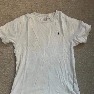Vit t-shirt från Polo Ralph Lauren - Vit t-shirt från Polo Ralph Lauren med klassisk rund hals och broderad marinblå logga på bröstet. Tillverkad i mjuk bomull, perfekt för en clean och stilren look. Passar dig som gillar enkla och snygga basplagg. Den kan ha några slitningar så ni vet för jag har använt den länge.