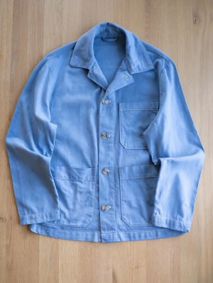 Ljusblå workwear Chore Jacket, size L - Snygg ljusblå workwear jacka i bomull med tre stora utanpåfickor och klassisk krage. Jackan har raka ärmar och stängs med fyra knappar framtill. Perfekt för en avslappnad och trendig stil. Tror Vintage HM