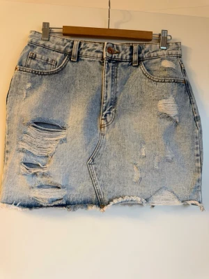 Ljusblå jeanskjol med slitningar - Trendig ljusblå jeanskjol i minilängd med rå kant och flera slitningar framtill. Kjolen har klassisk femficksmodell, bälteshällor och dragkedja med knapp. 