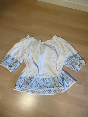 Vit blus med blått mönster - Säljer en vit offshoulder blus med ljusblått broderat mönster längs ärmar och nederkant. Blusen har trekvartsärm och resår vid axlarna för en avslappnad look. Perfekt till jeans eller kjol för en bohemisk vibe.