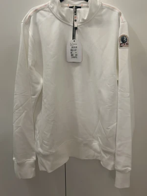 Vit halvzip hoodie från Parajumpers - Vit hoodie från Parajumpers med halv dragkedja framtill och broderad logga på vänster ärm. Tröjan har långa ärmar, ribbade muddar och orangea sömdetaljer vid dragkedjan. Perfekt för en clean och sportig look.