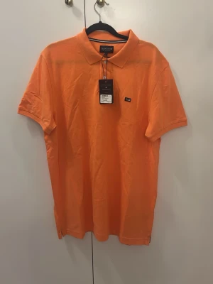 Orange pikétröja från Lexington - Snygg orange pikétröja från Lexington med klassisk krage och knappar framtill. Tröjan har en liten Lexington-logga på bröstet och är tillverkad i bomull. Perfekt för en avslappnad och stilren look.