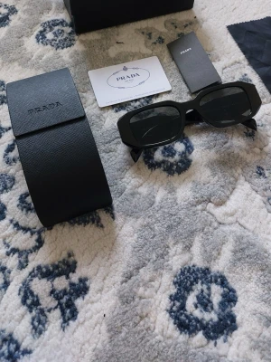 Svarta solglasögon från Prada - Snygga svarta solglasögon från Prada med rektangulära bågar och stilren design. Kommer med originalfodral och certifikat. Perfekta för dig som vill ha en trendig accessoar med lyxig känsla.