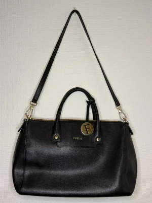 Svart handväska från Furla - Stilren svart handväska från Furla i slätt läder med guldfärgade detaljer och avtagbar axelrem. Väskan har två handtag, dragkedja upptill och en rund guldfärgad Furla-tag. Perfekt för dig som gillar klassisk och elegant design.