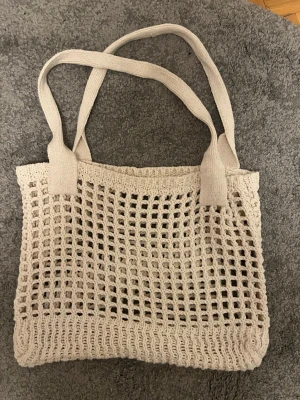 Beige virkad shoppingväska - Trendig shoppingväska i beige virkat material med rutigt nätmönster och breda axelband. Perfekt att slänga över axeln när du ska ut på stan eller till stranden. Rymmer allt du behöver och ger en chill vibe till din outfit.