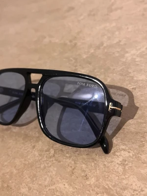 Svarta Tom Ford pilotbågar solglasögon - Snygga solglasögon från Tom Ford med klassisk pilotform. Svart båge i syntetmaterial och blåtonade glas. Guldiga detaljer vid gångjärnen ger en exklusiv touch. Perfekta för dig som vill ha en stilren och trendig accessoar.