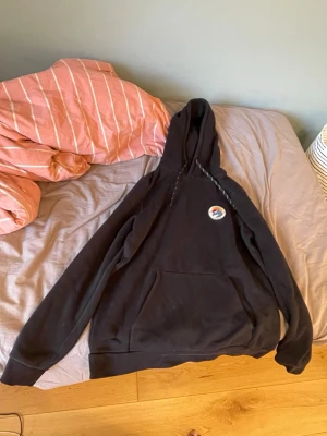 Svart hoodie med liten patch - Säljer en svart hoodie med huva och känguruficka framtill. På bröstet sitter en liten rund patch med färgglatt motiv. Hoodien har långa ärmar och ribbade muddar. Perfekt för dig som gillar enkel och stilren streetwear.
