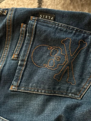 Philipp Plein jeans ( vintage äkta )  - Säljer ett par blå jeans från Philipp Plein med cool dödskalle och korslagda ben broderat på bakfickan. Klassisk femficksmodell med kontrastsömmar och en edgy streetwear-vibe. Perfekta för dig som vill sticka ut lite extra.