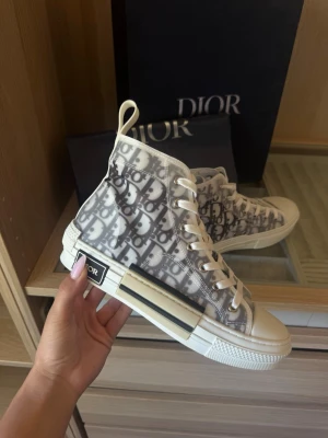Dior B23 high-top sneakers - Snygga Dior B23 high-top sneakers med genomskinligt tyg och ikoniskt Dior-monogram i grått och vitt. Skorna har vita snören, gummisula med svarta och vita detaljer samt en modern siluett. Perfekta för dig som vill sticka ut med exklusiv streetstyle.