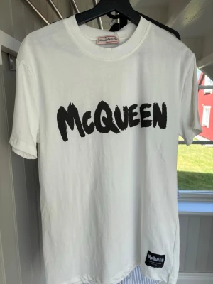 Vit McQueen t-shirt i bomull - Snygg vit t-shirt från Alexander McQueen med svart tryck framtill och en liten patch nertill. Klassisk rund halsringning och korta ärmar. Tillverkad i mjuk bomull för en skön känsla och stilren look.