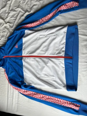 Blå och vit vindjacka från Le Coq Sportif - Retroinspirerad vindjacka i blått och vitt från Le Coq Sportif. Jackan har röda och vita rutiga detaljer längs ärmarna, hög krage och röda dragkedjor. Tillverkad i polyester med två fickor framtill och ribbade muddar.