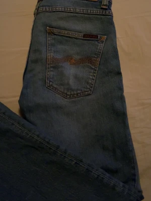 Blå jeans från Nudie Jeans - Säljer ett par klassiska blå jeans från Nudie Jeans med snyggt slitna detaljer och kontrastsömmar. Jeansen har fem fickor, knappgylf och en cool broderad bakficka. Perfekta för en avslappnad och trendig look. Dom är uppsydda så lite kortare i storleken!