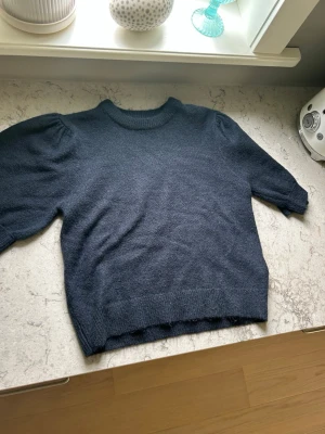 Mörkblå stickad tröja Gina Tricot - Mysig mörkblå stickad tröja från Gina Tricot med puffärmar och ribbade muddar vid hals, ärmslut och nederkant. Tröjan har en klassisk rund halsringning och är perfekt för lager på lager under kyliga dagar.