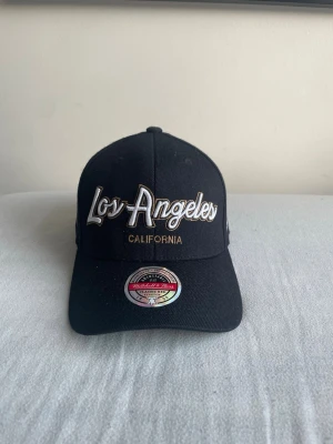 Svart Los Angeles keps - Snygg svart keps från Mitchell & Ness med broderad text 'Los Angeles California' i vitt och guld på framsidan. Klassisk böjd skärm och justerbar baksida. Perfekt accessoar för dig som gillar streetwear och sportig stil.