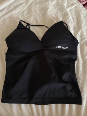 dfyne top - Snygg svart crop top från DFYNE med tunna axelband och logga på bröstet. Toppen har en figurnära passform och är tillverkad i ett stretchigt material som passar perfekt till träning eller som ett trendigt basplagg.