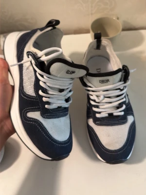 Dior sneakers i blått och vitt - Snygga Dior sneakers i blått och vitt med meshpaneler och mockadetaljer. Klassisk låg modell med vita skosnören och tydlig Dior-logga på plösen. Perfekta för dig som vill ha en clean och exklusiv look till vardagsoutfiten.