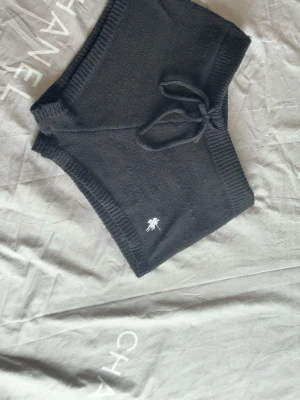 Svarta stickade shorts med snörning - Svarta stickade shorts med hög midja och snörning i midjan. Shortsens material är mjukt och ribbstickat, och de har en liten broderad detalj framtill. Perfekta för en avslappnad och trendig look.