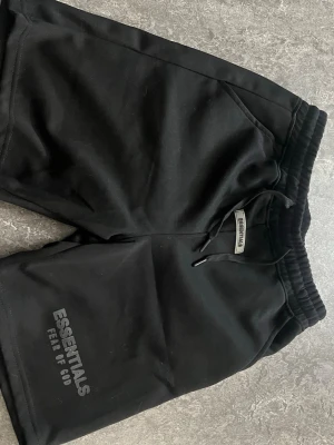 Essentials Shorts - Säljer ett par svarta shorts från Essentials Fear of God med snörning i midjan och tryckt logga på benet. Shortsen har en relaxed passform och är tillverkade i mjukt material, perfekta för chill dagar.