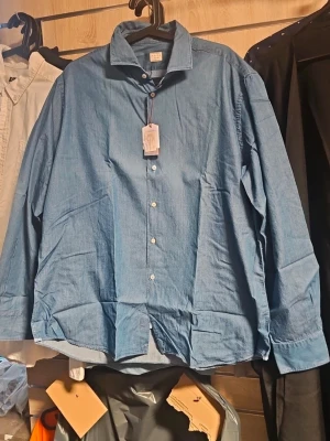 Blå skjorta från xakus i denim - Snygg blå skjorta från Nacus i mjuk bomull. Klassisk krage, knäppning framtill med vita knappar och lång ärm. Perfekt för en clean och avslappnad stil.