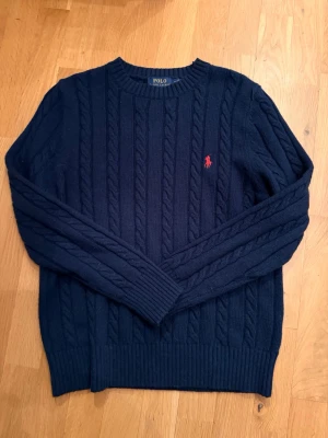 Mörkblå kabelstickad tröja Polo Ralph Lauren - Kabelstickad mörkblå tröja från Polo Ralph Lauren med rund halsringning och röd broderad logga på bröstet. Tröjan har ribbade muddar vid ärmslut och nederkant, perfekt för en klassisk och stilren look. Säljer då den var för lite för mig priset kan diskuteras då jag inte har någon användning av den hör av er vid funderingar den är helt oanvänd 