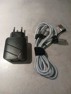 Laddare med multikabel USB och adapter - Säljer en svart väggadapter med två USB-portar och en silverfärgad flätad multikabel. Kabeln har USB-A i ena änden och tre olika kontakter (Micro-USB, USB-C och Lightning) i andra änden. Perfekt för dig som vill ladda flera olika enheter med samma kabel.