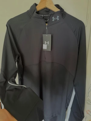 Svart träningströja från Under Armour - Snygg svart långärmad träningströja från Under Armour med halv dragkedja framtill och hög krage. Tillverkad i ett stretchigt funktionsmaterial som andas bra, perfekt för löpning eller gym. Diskret logga på bröstet och stilren design.