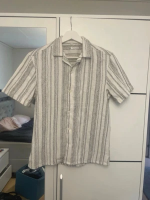 Randig kortärmad skjorta från Zara - Säljer en snygg kortärmad skjorta från Zara i vitt och grått med vertikala ränder. Skjortan har klassisk krage, knappar framtill och är tillverkad i ett luftigt material som passar perfekt till varma dagar.