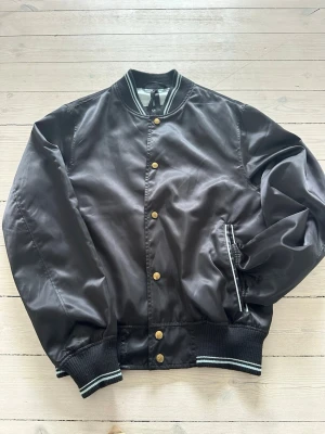 Svart bomberjacka  - Snygg svart bomberjacka med ribbade muddar och krage med vita ränder. Jackan har guldfärgade knappar framtill och två fickor med randiga detaljer. Klassisk passform och sportig look, perfekt till streetwear-stilen.